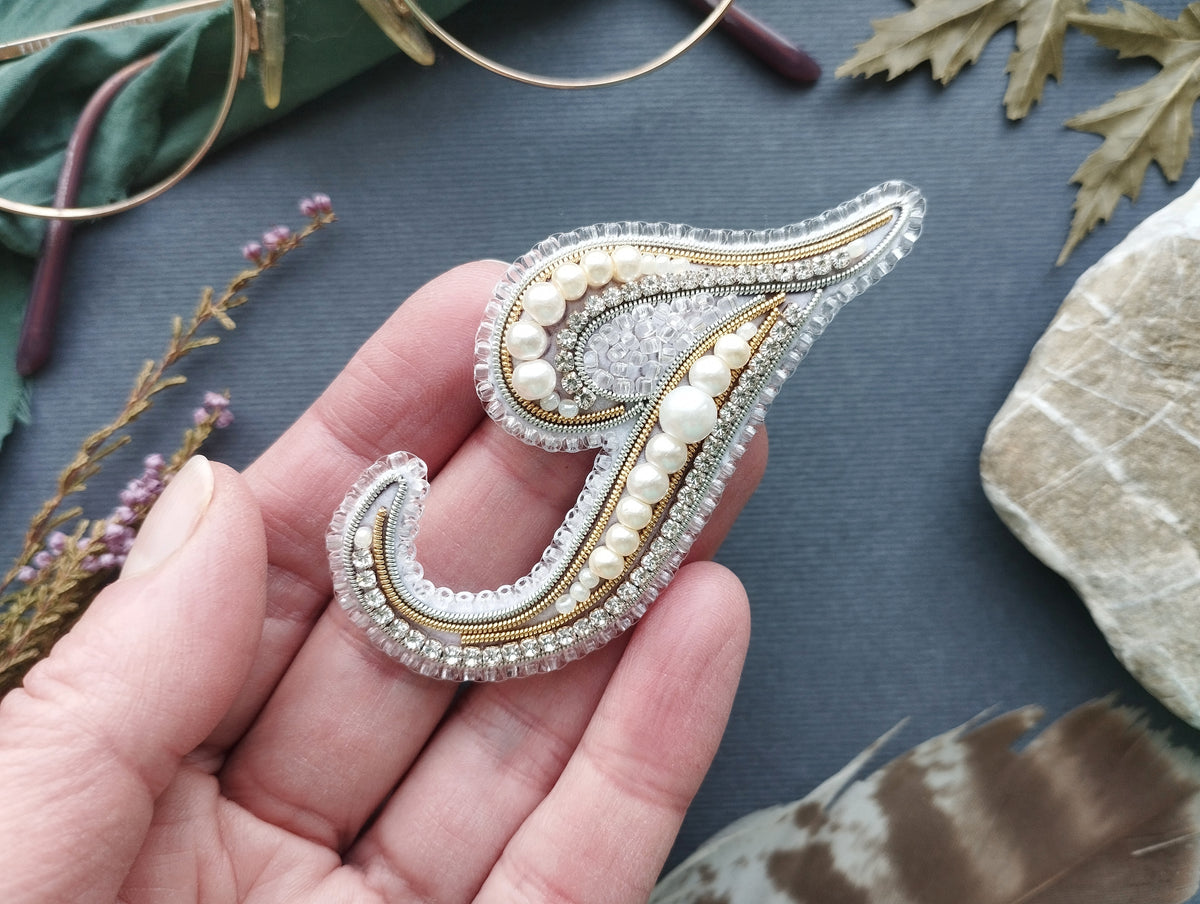 A hand holding a decorative brooch with pearls and a metallic design, surrounded by various natural elements such as leaves and feathers.