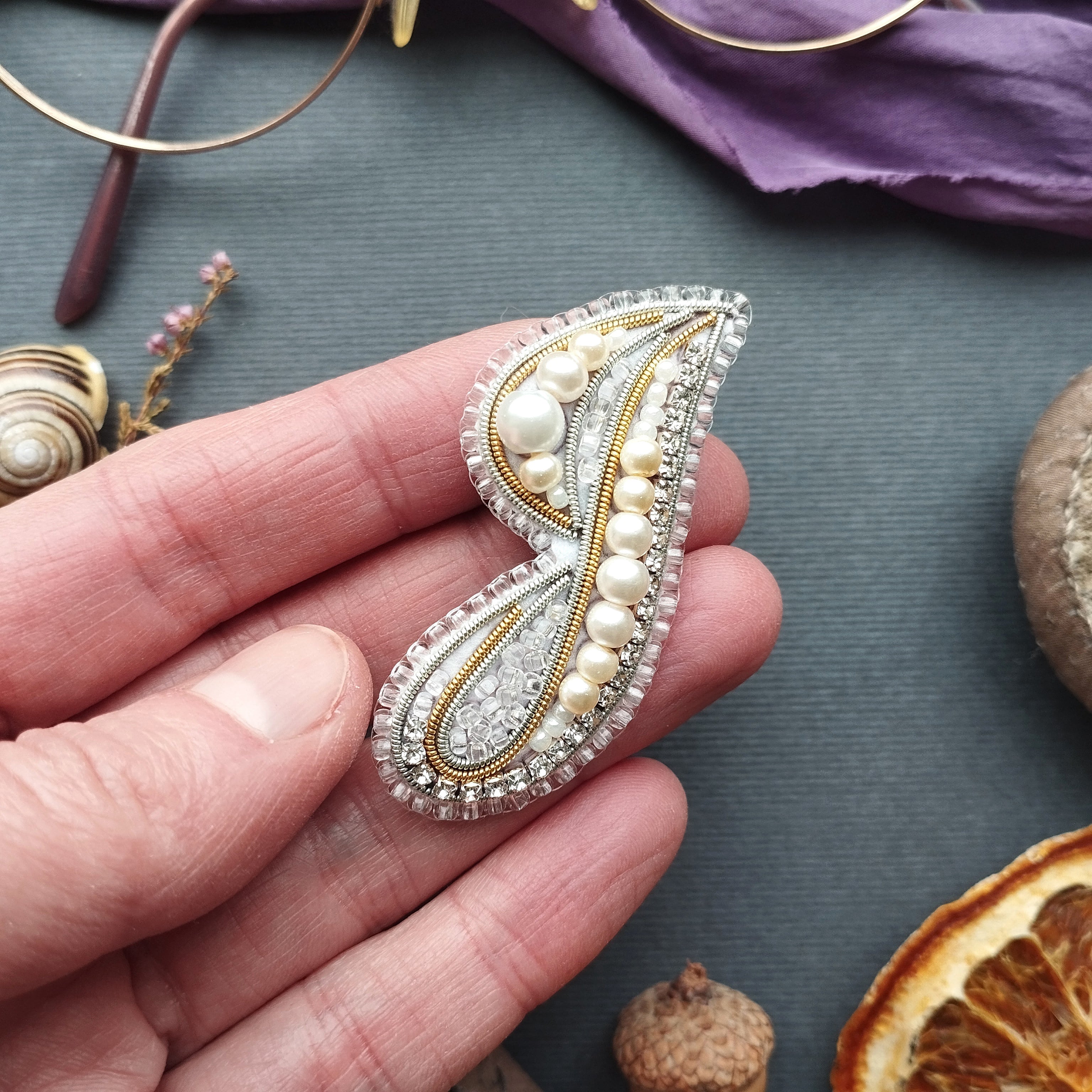 A hand holding a decorative piece of jewelry, such as a brooch or pendant, against a backdrop of various objects including a pair of glasses, a purple fabric, and dried fruit.