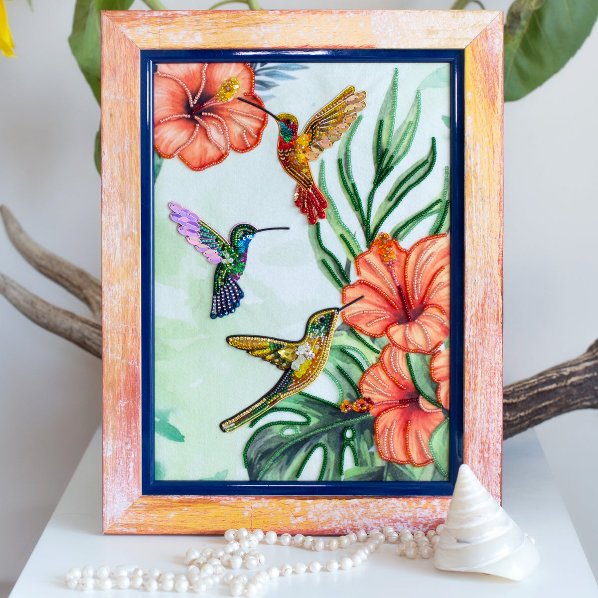 a framed painting of three hummingbirds in flight, surrounded by vibrant flowers and lush foliage.
