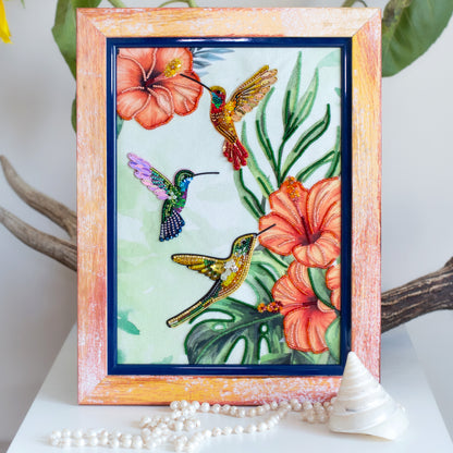 a framed painting of three hummingbirds in flight, surrounded by vibrant flowers and lush foliage.
