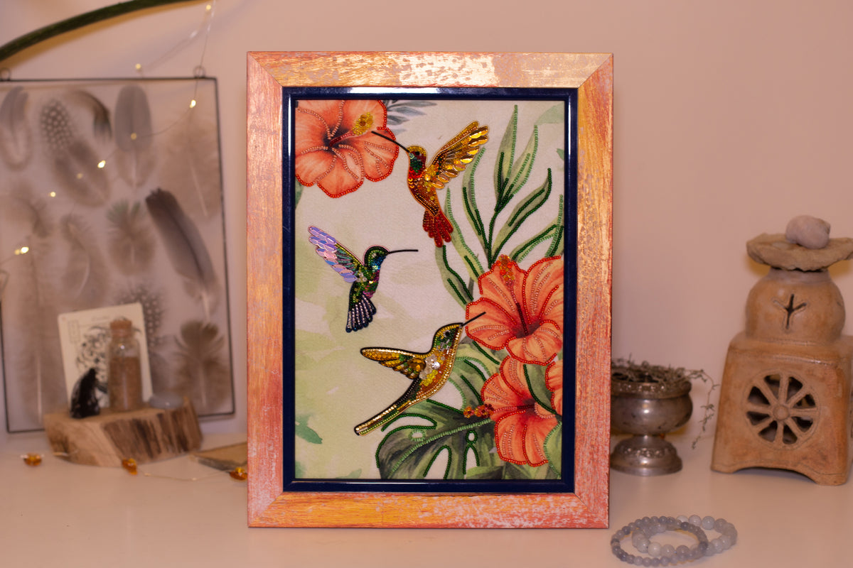 a framed painting of three hummingbirds hovering over a vibrant floral arrangement, with a wooden frame and a warm, earthy color palette.