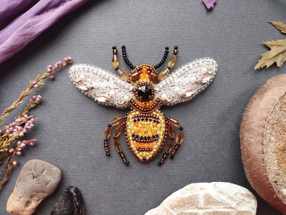 a detailed and intricately designed beaded bee pin, surrounded by various natural elements such as leaves, flowers, and rocks.