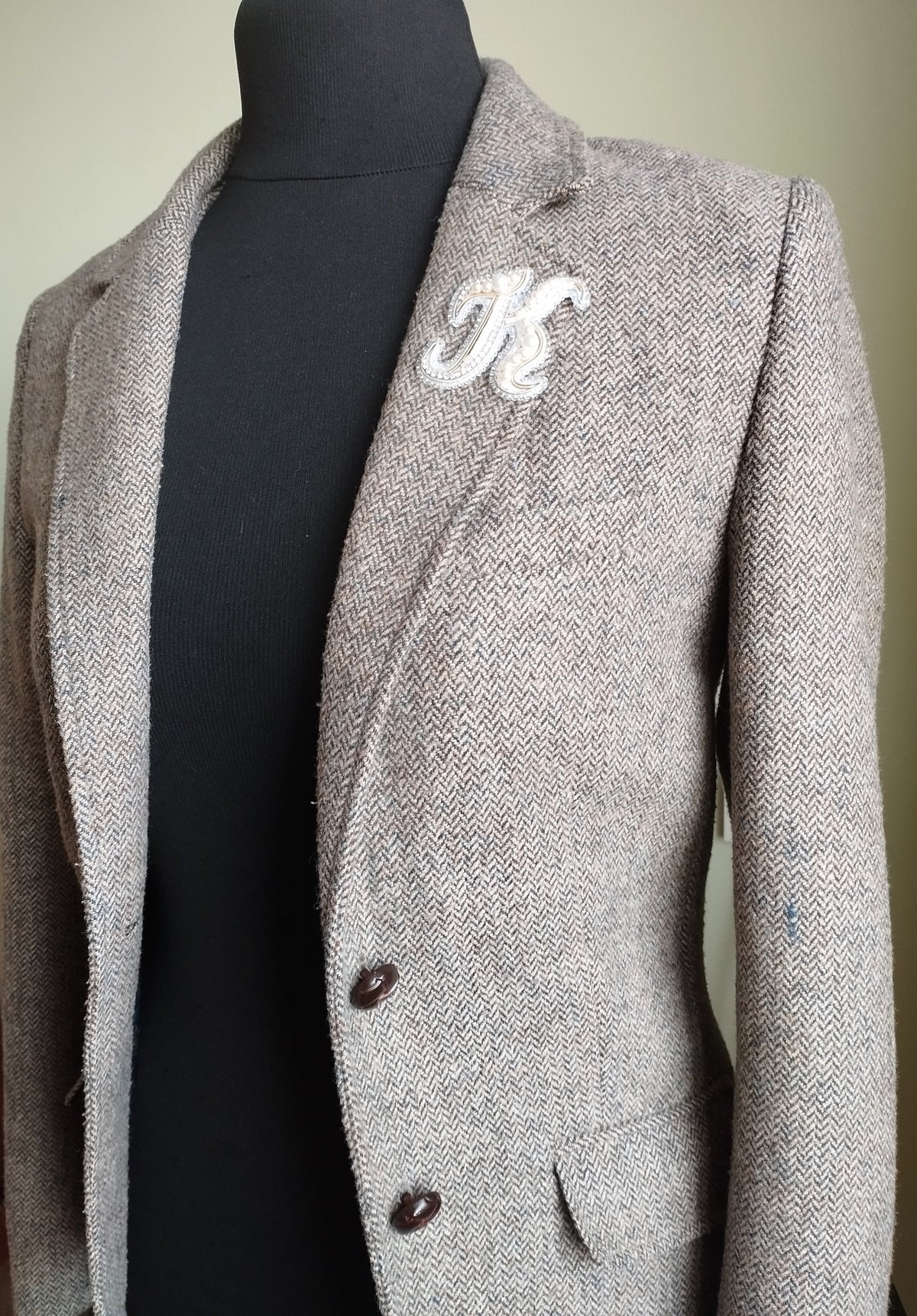 A gray tweed blazer with a white "K" embroidered on the left side, displayed on a mannequin.