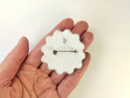 A hand holding a white fabric pin with a silver pin attached to it.