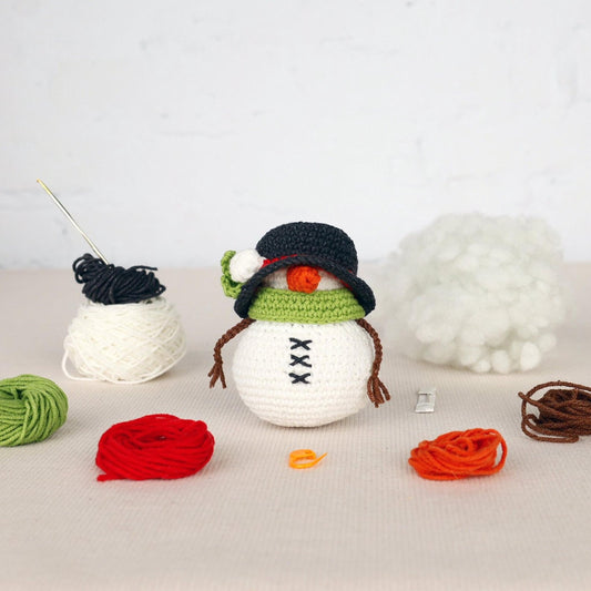 A crocheted snowman figurine surrounded by various crochet yarns and a crochet hook, with a pile of white pom-poms in the background.