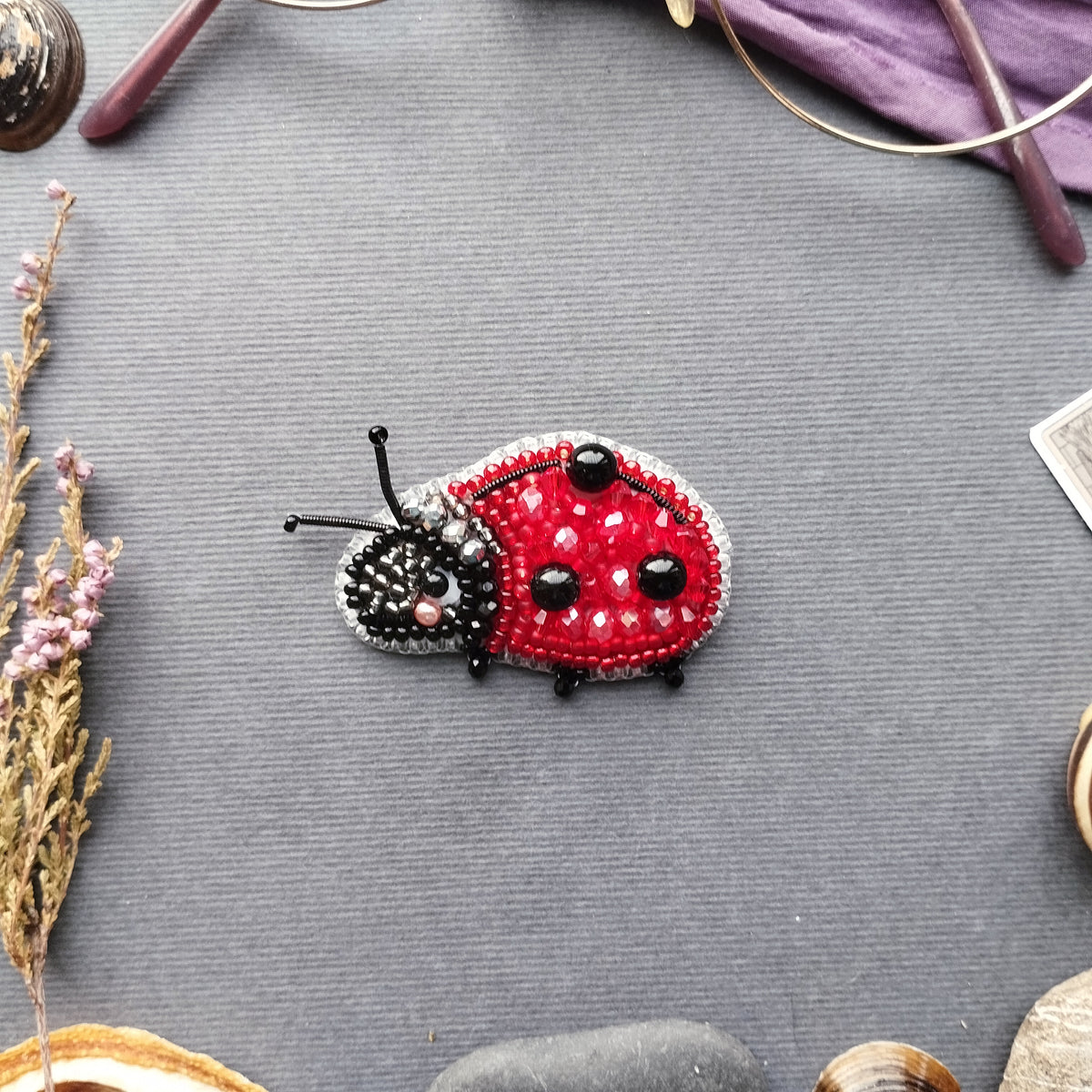 A red and black beaded ladybug brooch is displayed on a gray surface, surrounded by various objects such as a magnifying glass, a flower, and a rock.