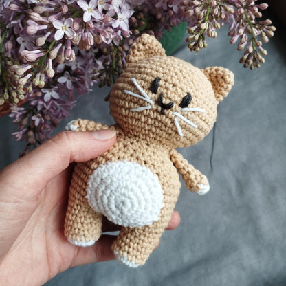 a hand holding a small crocheted cat next to a bouquet of flowers