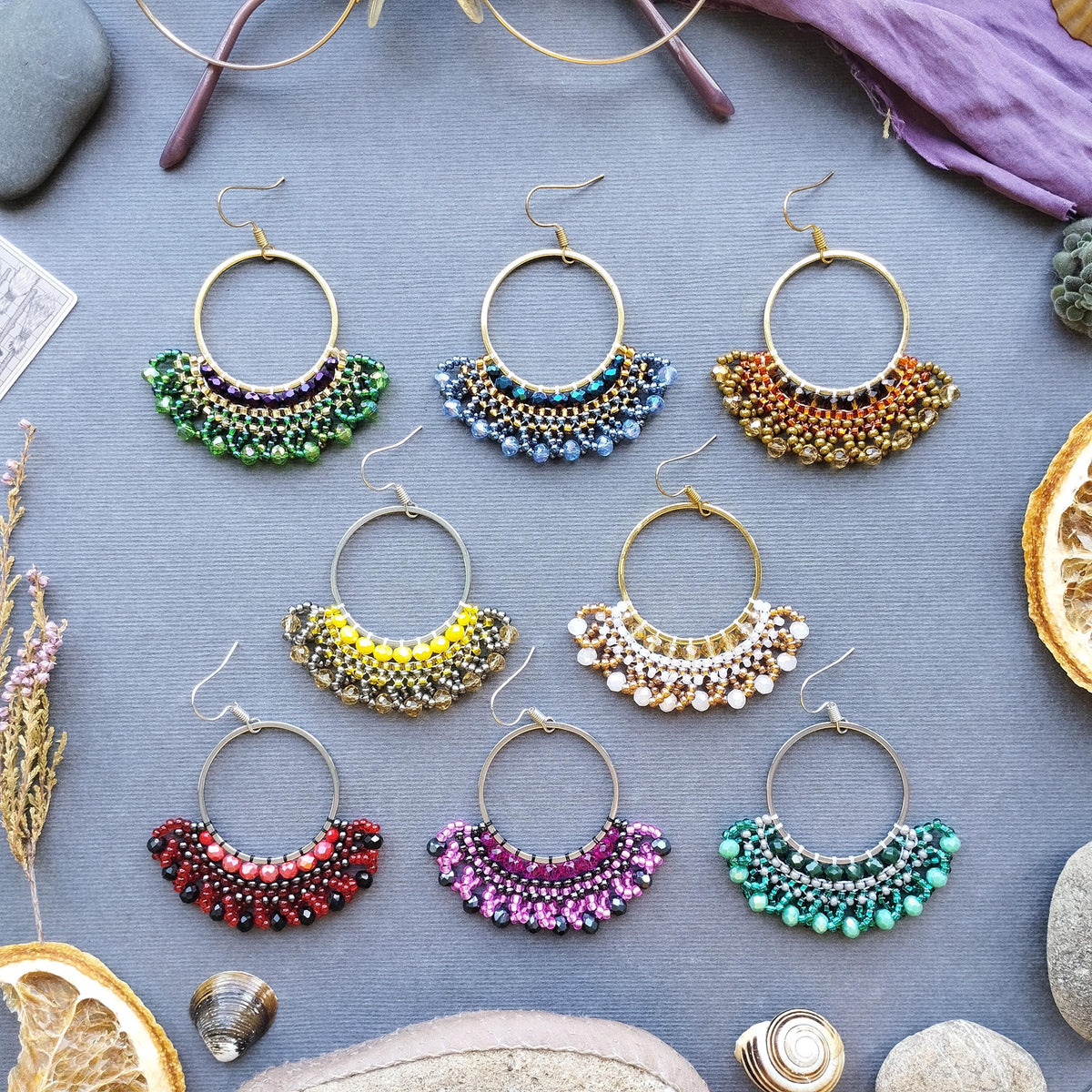 A collection of colorful beaded earrings in various shapes and sizes, arranged on a gray background with some dried flowers and fruits.