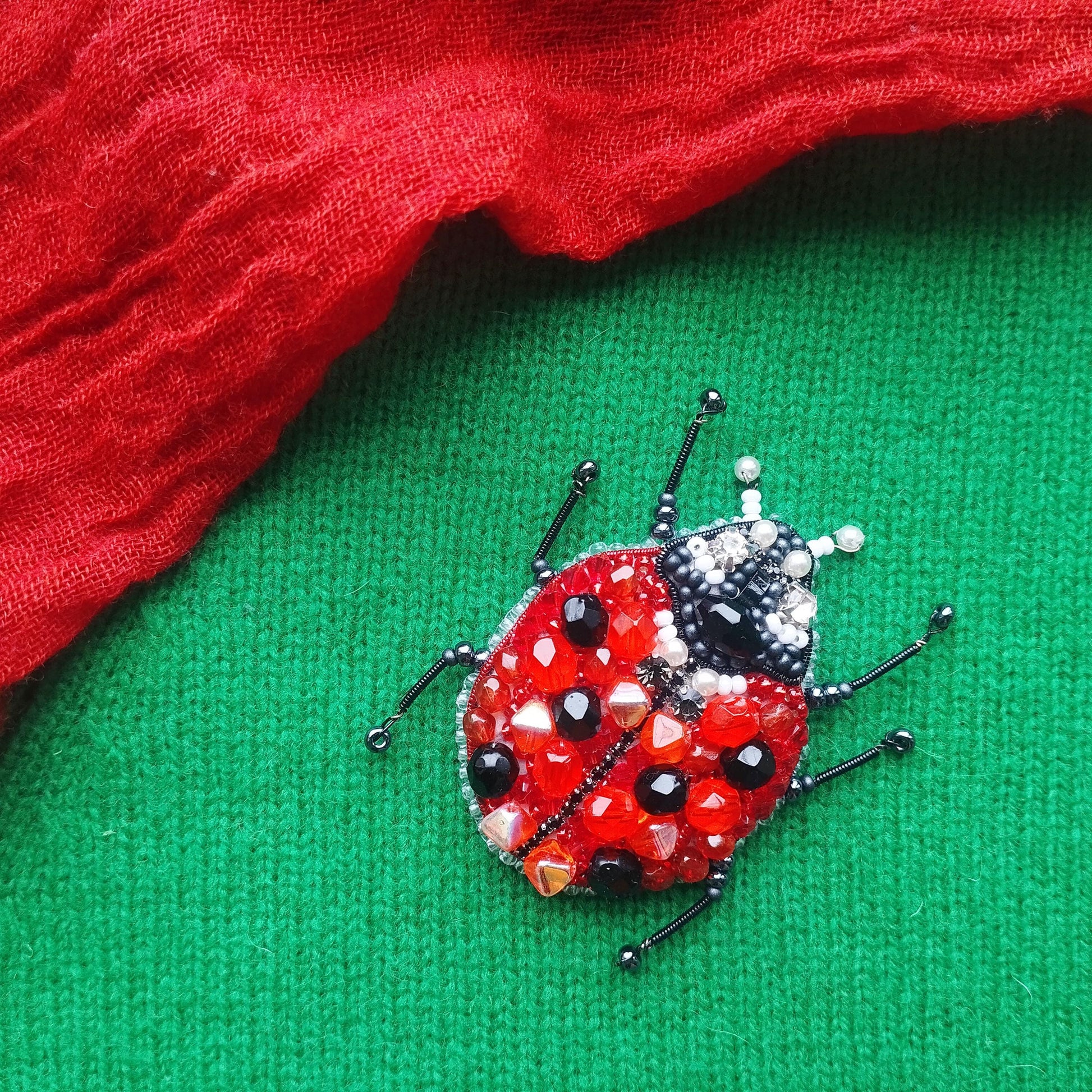 A red and black ladybug brooch with beaded details is displayed on a green fabric surface.