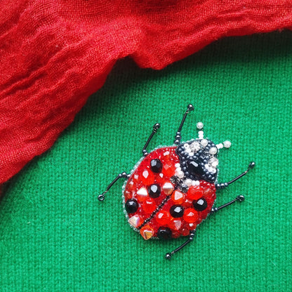 A red and black ladybug brooch with beaded details is displayed on a green fabric surface.