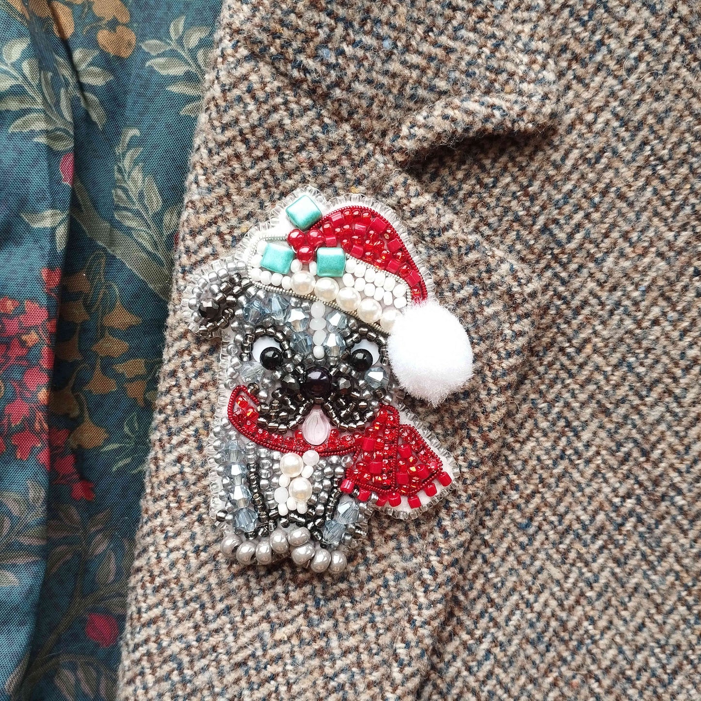 A close-up of a decorative button or pin featuring a small dog wearing a Santa hat and scarf, with a floral patterned fabric visible in the background.