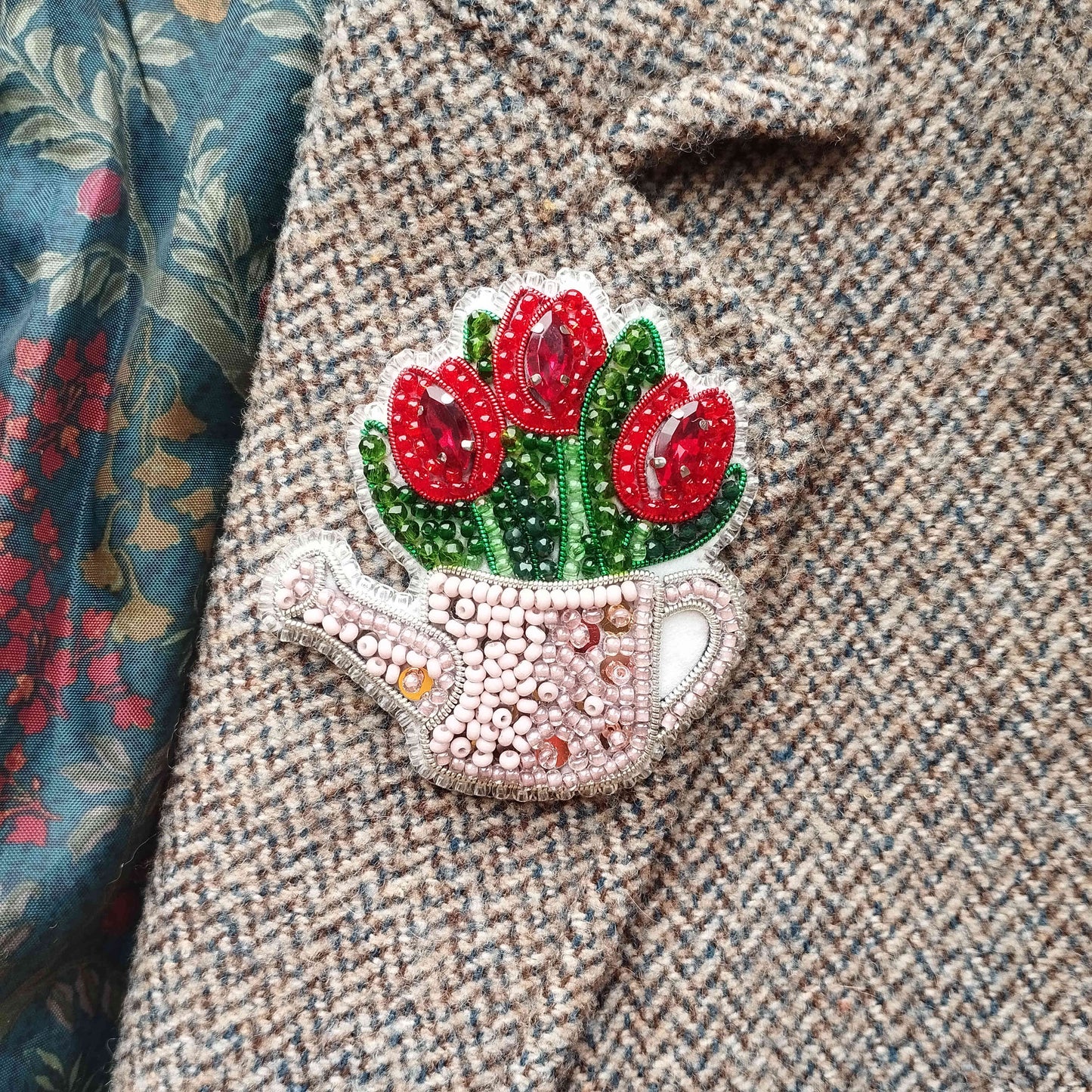 A close-up of a beaded, embroidered pin or brooch on a tweed jacket, featuring a design of red flowers.
