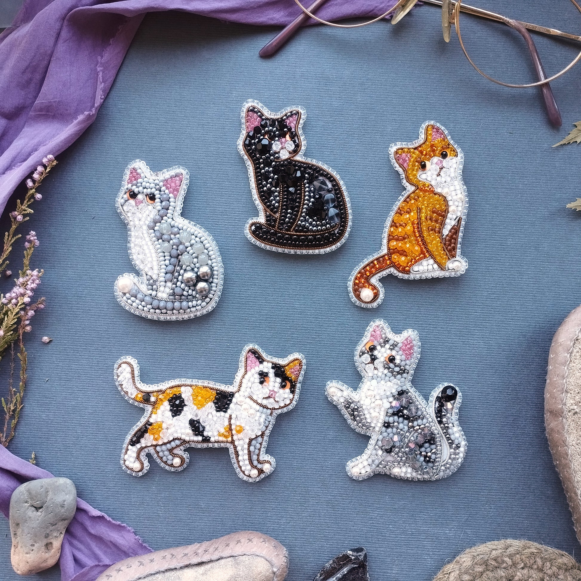 six beaded cat brooches arranged on a blue background, with some of the cats depicted in different colors and poses.
