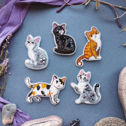 six beaded cat brooches arranged on a blue background, with some of the cats depicted in different colors and poses.