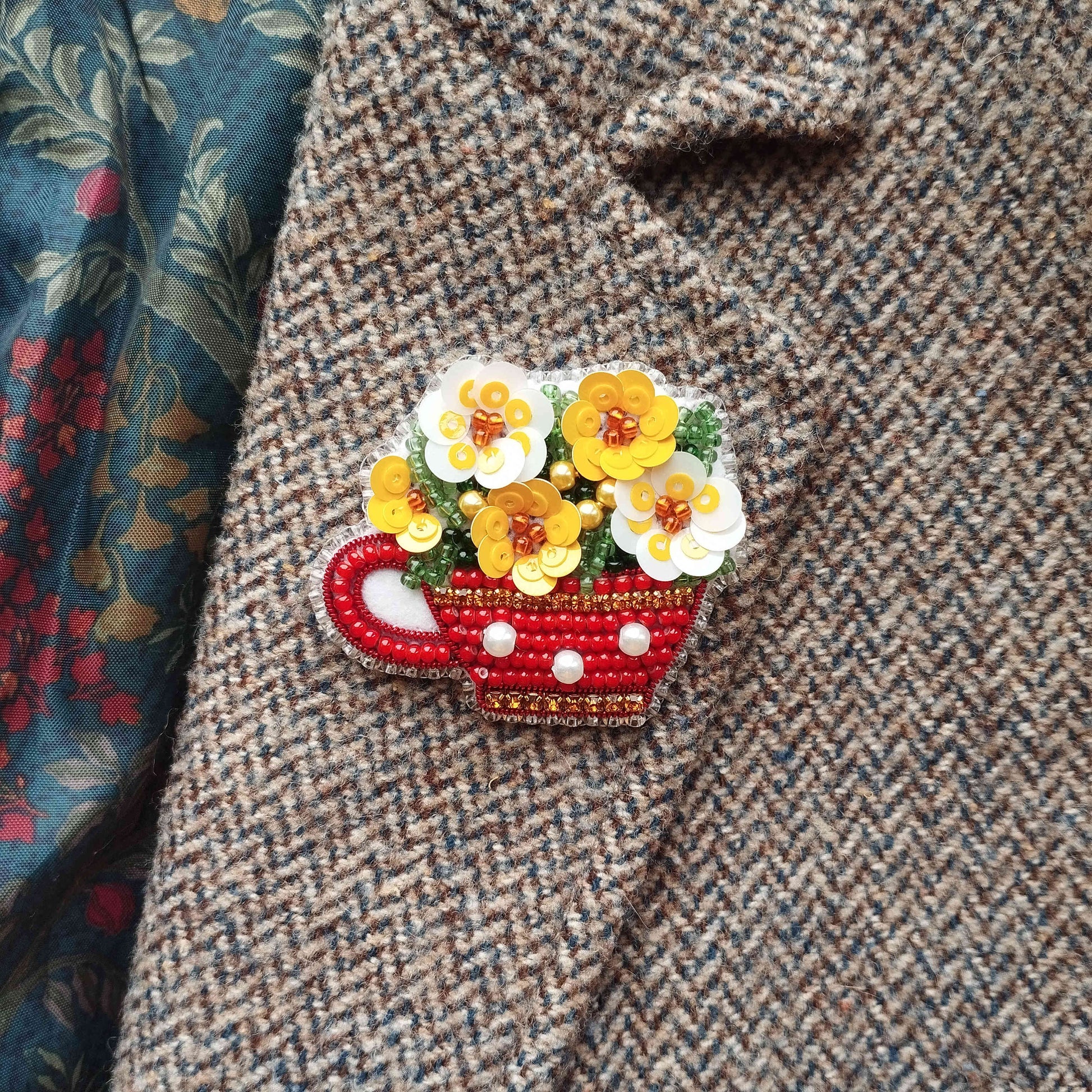 A close-up of a red and gold brooch with yellow flowers attached to a grey tweed jacket.
