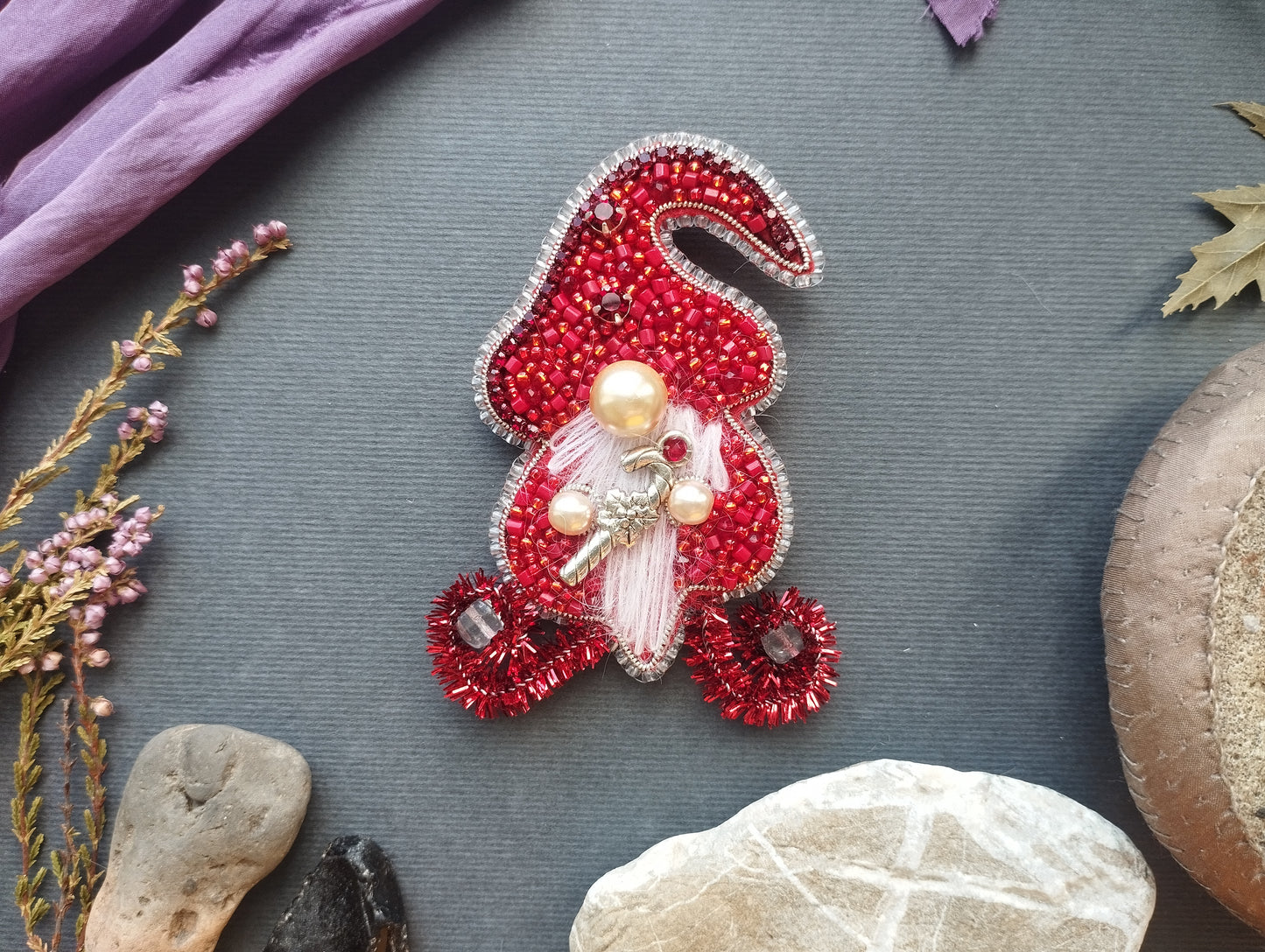 a red ornament with a gnome-like design, adorned with beads and pearls, placed on a gray surface. Surrounding the ornament are various natural elements, including a purple fabric, a small plant, and some rocks.
