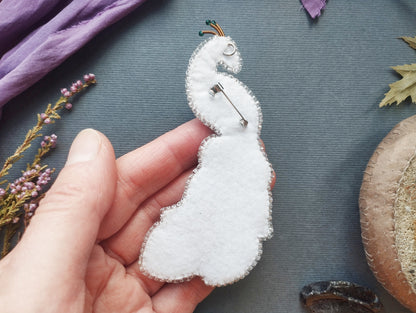 A hand holding a white felt peacock brooch against a blue background with various natural elements, such as leaves and flowers.