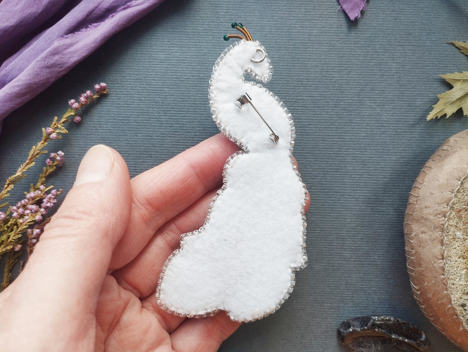 A hand holding a white felt peacock brooch against a blue background with various natural elements, such as leaves and flowers.