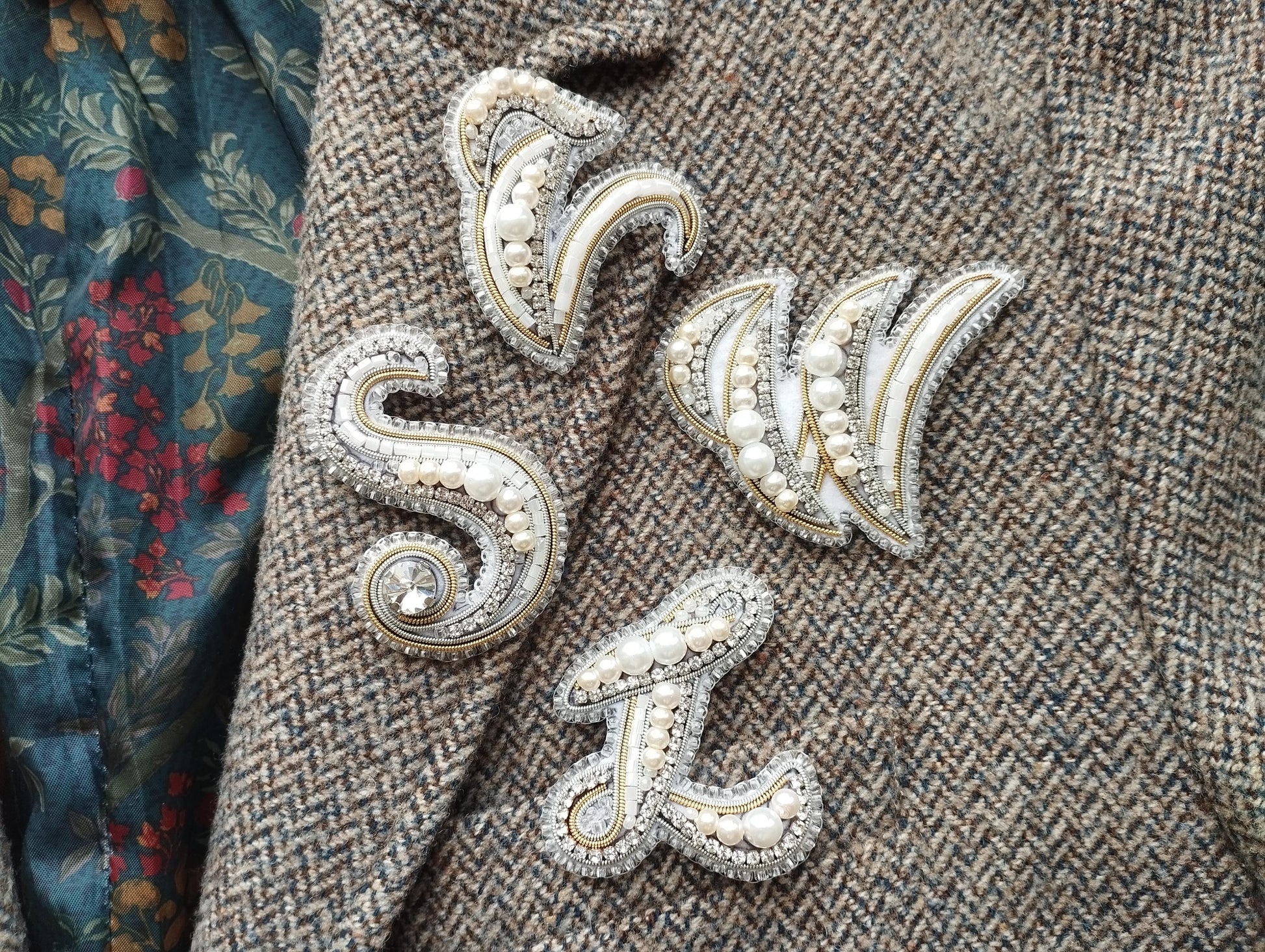 a close-up view of a grey tweed jacket with several decorative embellishments, including a large letter "S" and a smaller letter "W" made of pearls and rhinestones.