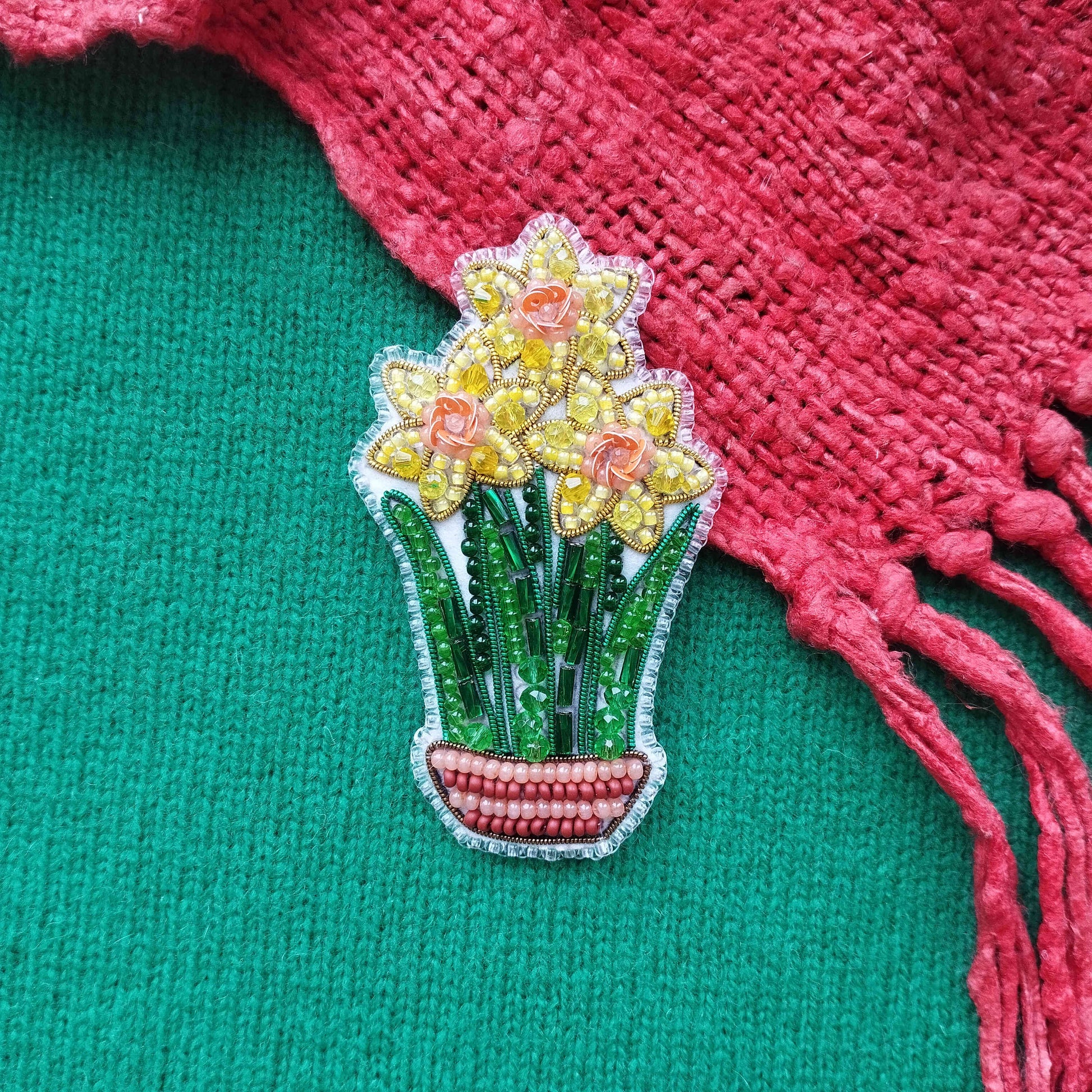 A decorative beaded brooch in the shape of a flower arrangement, featuring yellow flowers and green leaves, is displayed on a green fabric surface next to a red fabric fringe.