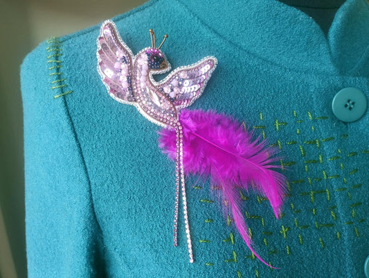 A close-up of a teal-colored jacket with a decorative pin featuring a pink bird-like design and a long pink feather attached to the right side of the jacket.