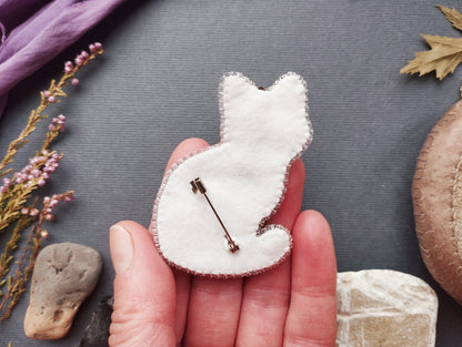 A hand holding a white felt cat pin against a gray background, with various natural elements such as leaves and rocks in the background.