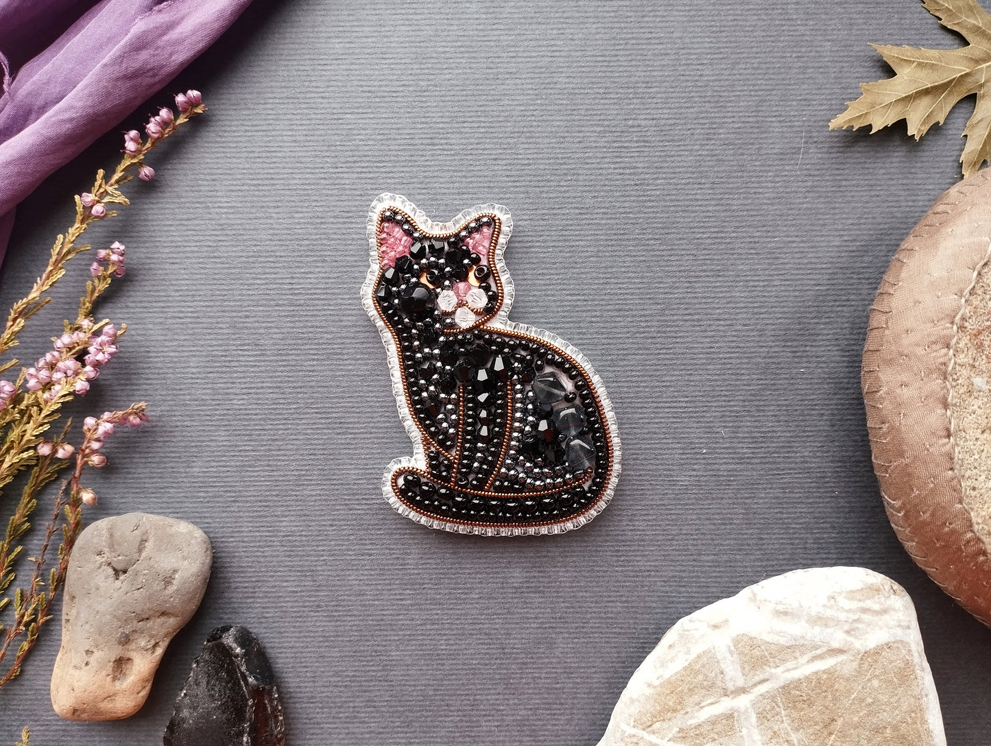 a black cat embroidered with beads and surrounded by various objects, including a purple fabric, a leaf, and some rocks.