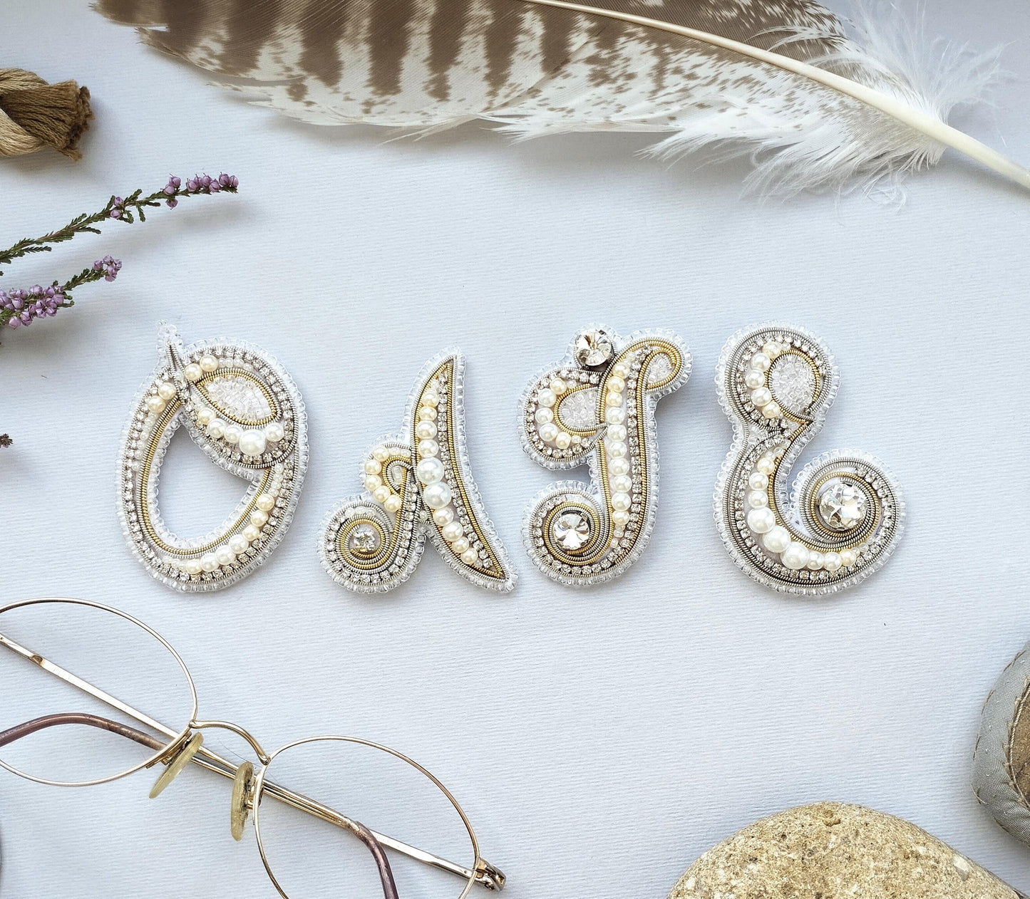 a close-up of a collection of ornate, bejeweled letters spelling out the word "O". The letters are arranged in a visually appealing manner, with the word "O" being the most prominent. The image also includes various other decorative elements, such as a feather, a pair of glasses, and some flowers, adding to the overall aesthetic of the scene.