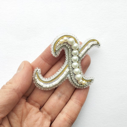 A hand holding a decorative letter 'X' made of beads, pearls, and other embellishments.