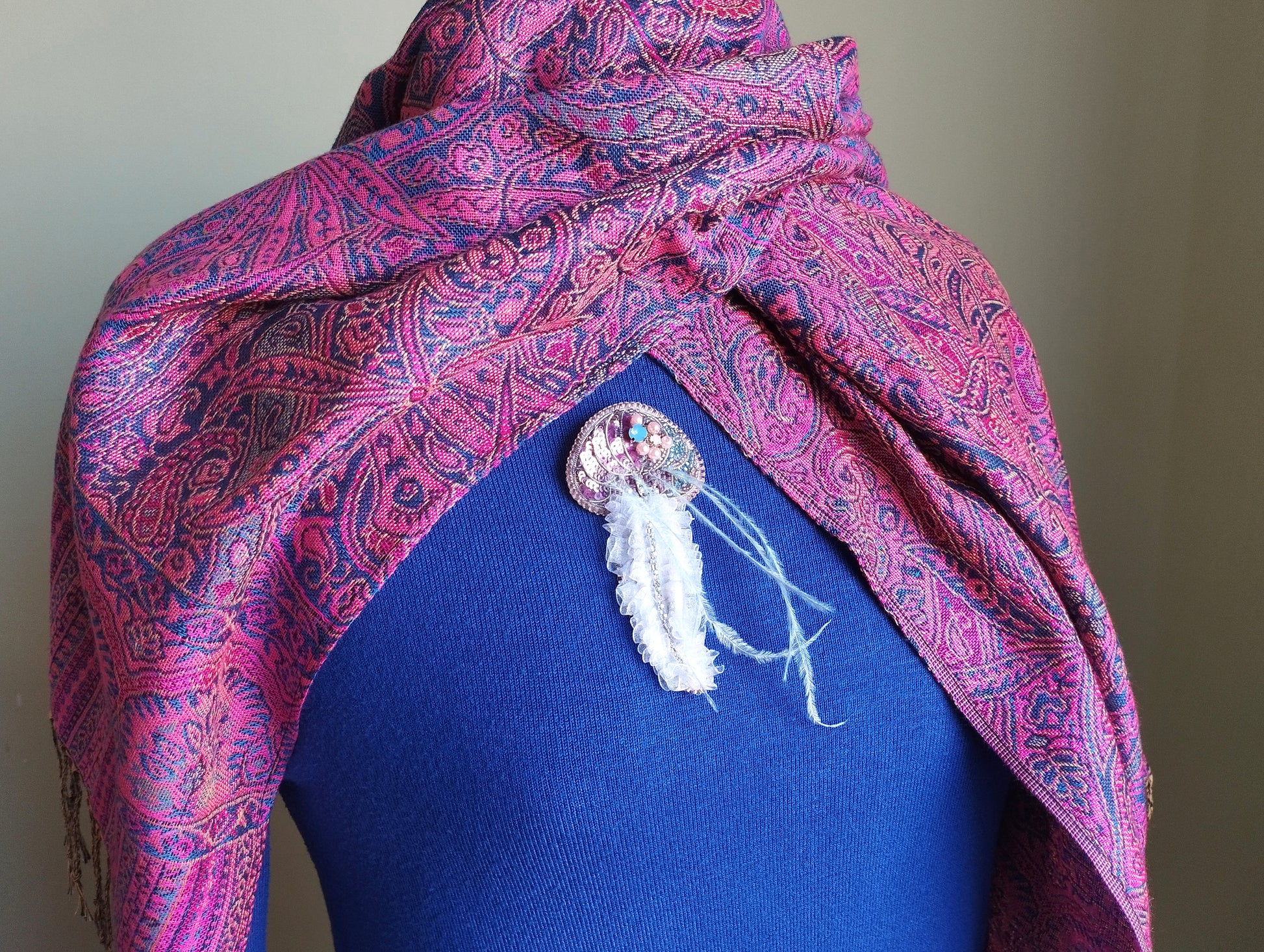 A mannequin wearing a blue top with a pink and purple paisley patterned scarf draped over it. The scarf has a white feather attached to it, and there is a small decorative pin or brooch on the chest area.