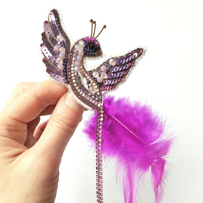 A hand is holding a decorative pin or brooch in the shape of a bird, with a feather attached to it.