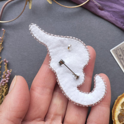 A hand holding a white felt elephant pin with a silver pin attached to it, surrounded by various objects such as a pair of glasses, a purple fabric, and a slice of orange.