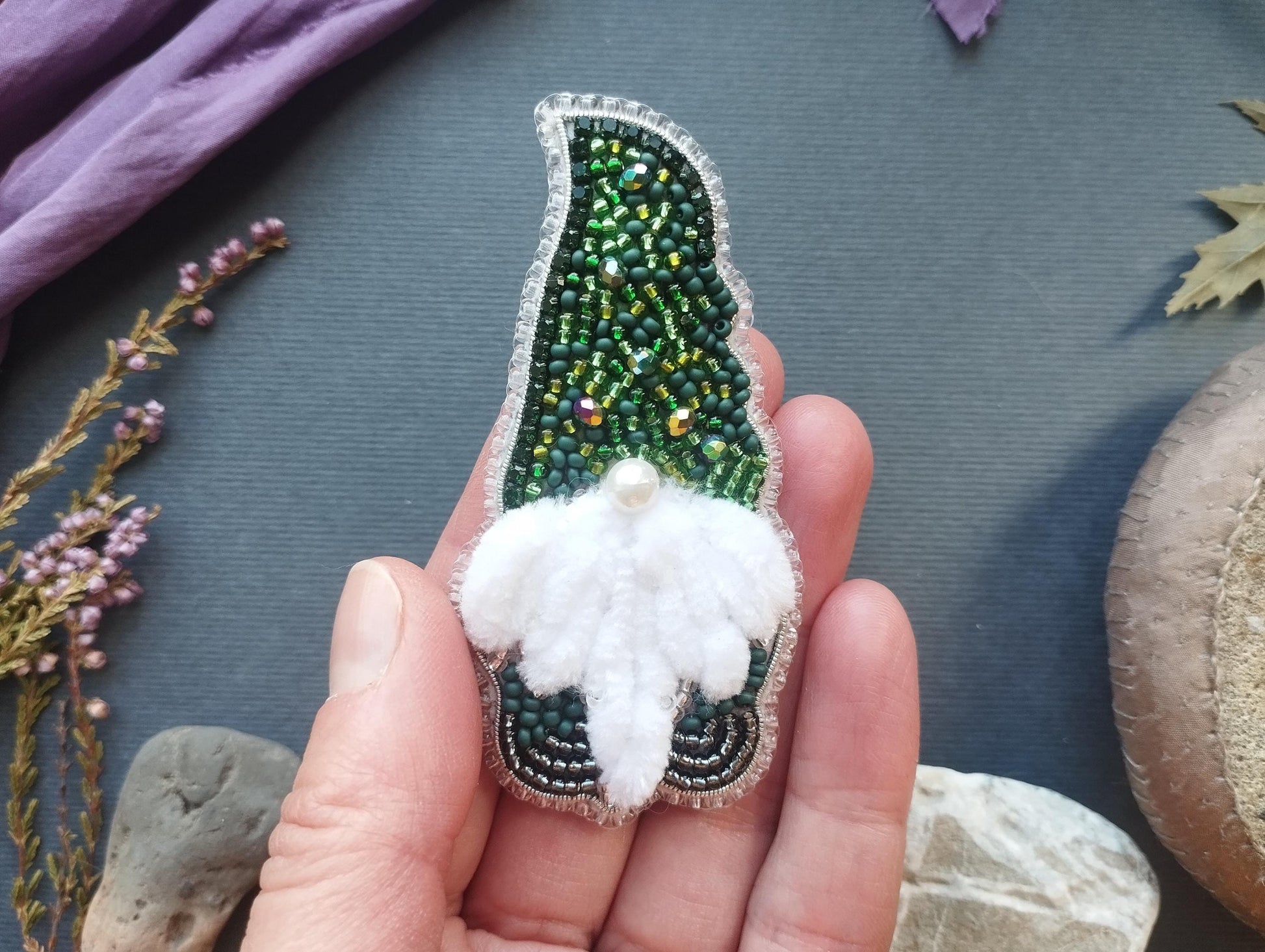 A hand holding a small, intricately designed ornament made of green and white beads, with a white flower-like design on the front.