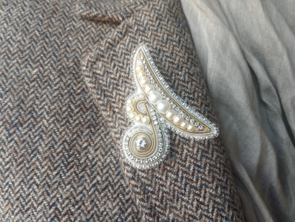 A close-up image of a decorative brooch or pin with pearls and a metallic design, attached to a tweed fabric.