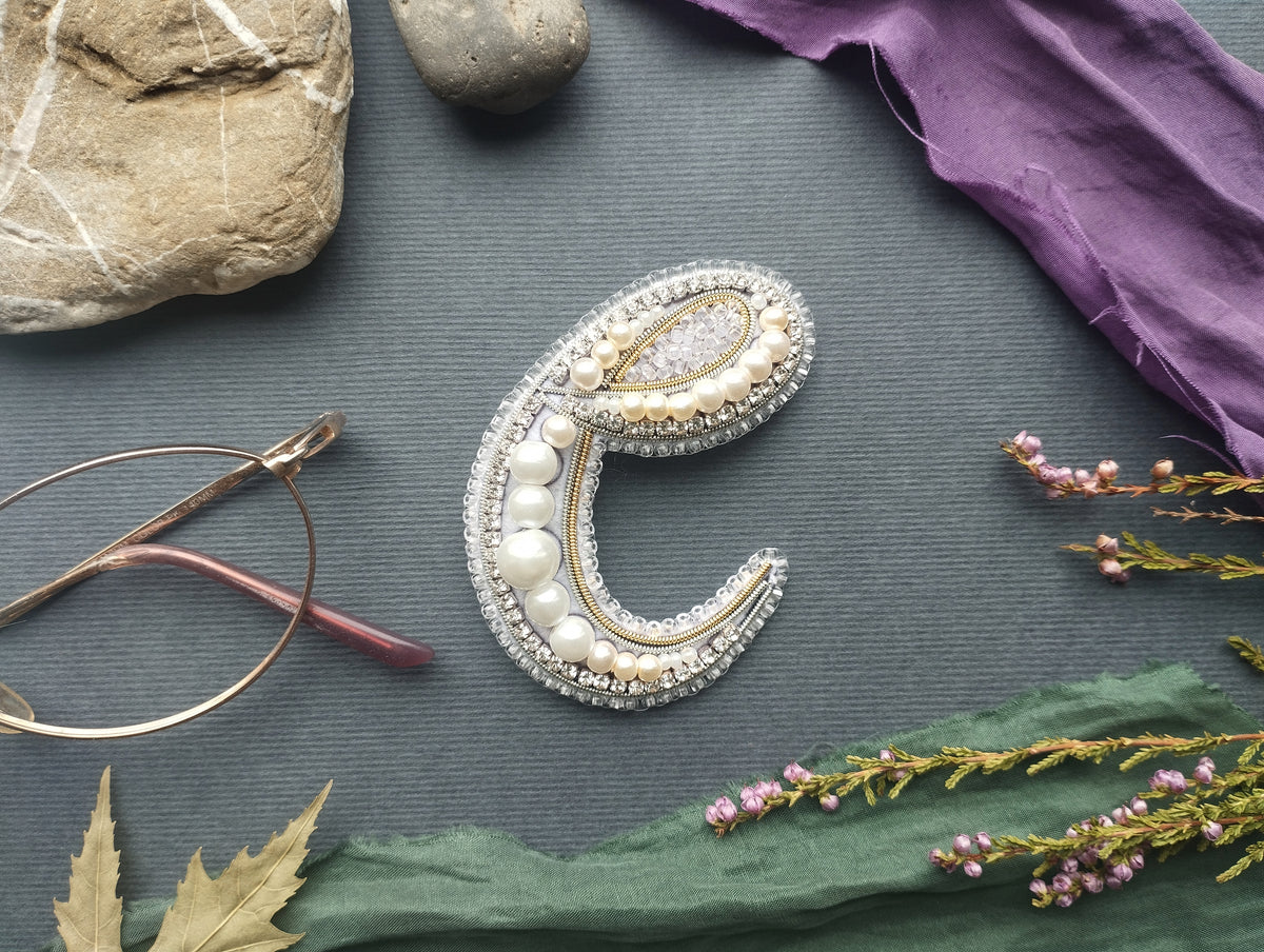 a close-up of a decorative letter 'C' made of beads, pearls, and other embellishments, surrounded by various objects such as a rock, a pair of glasses, and flowers.