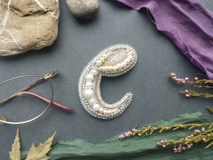 a close-up of a decorative letter 'C' made of beads, pearls, and other embellishments, surrounded by various objects such as a rock, a pair of glasses, and flowers.