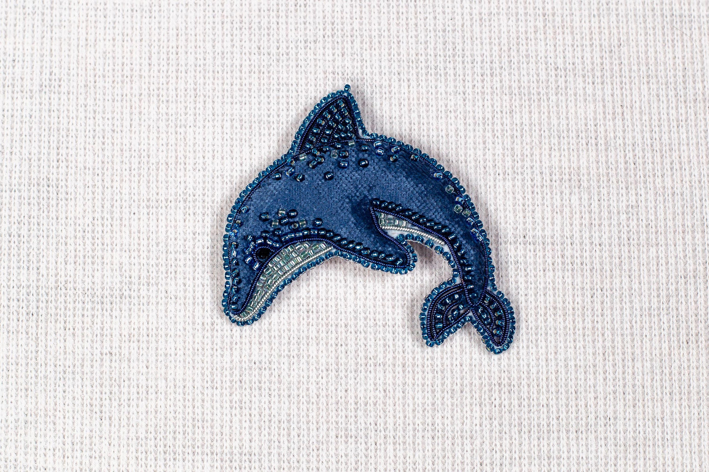 A blue, hand-beaded dolphin brooch is displayed against a white background.