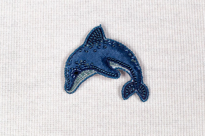 A blue, hand-beaded dolphin brooch is displayed against a white background.