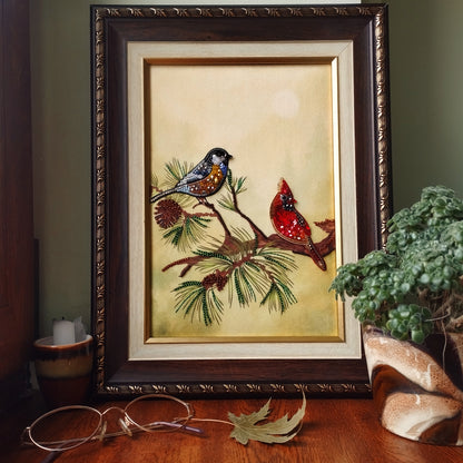 a framed painting of two birds perched on a branch, surrounded by pine cones and pine needles, placed on a wooden surface.