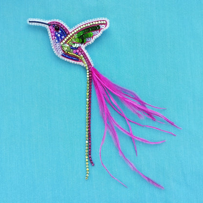 A colorful, intricately designed hummingbird brooch made of various materials, including jewels, feathers, and beads, is displayed against a bright blue background.