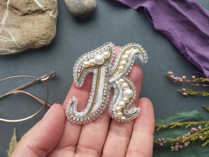 A hand holding a decorative letter "K" made of beads and pearls, surrounded by various objects such as a rock, a purple fabric, and some flowers.