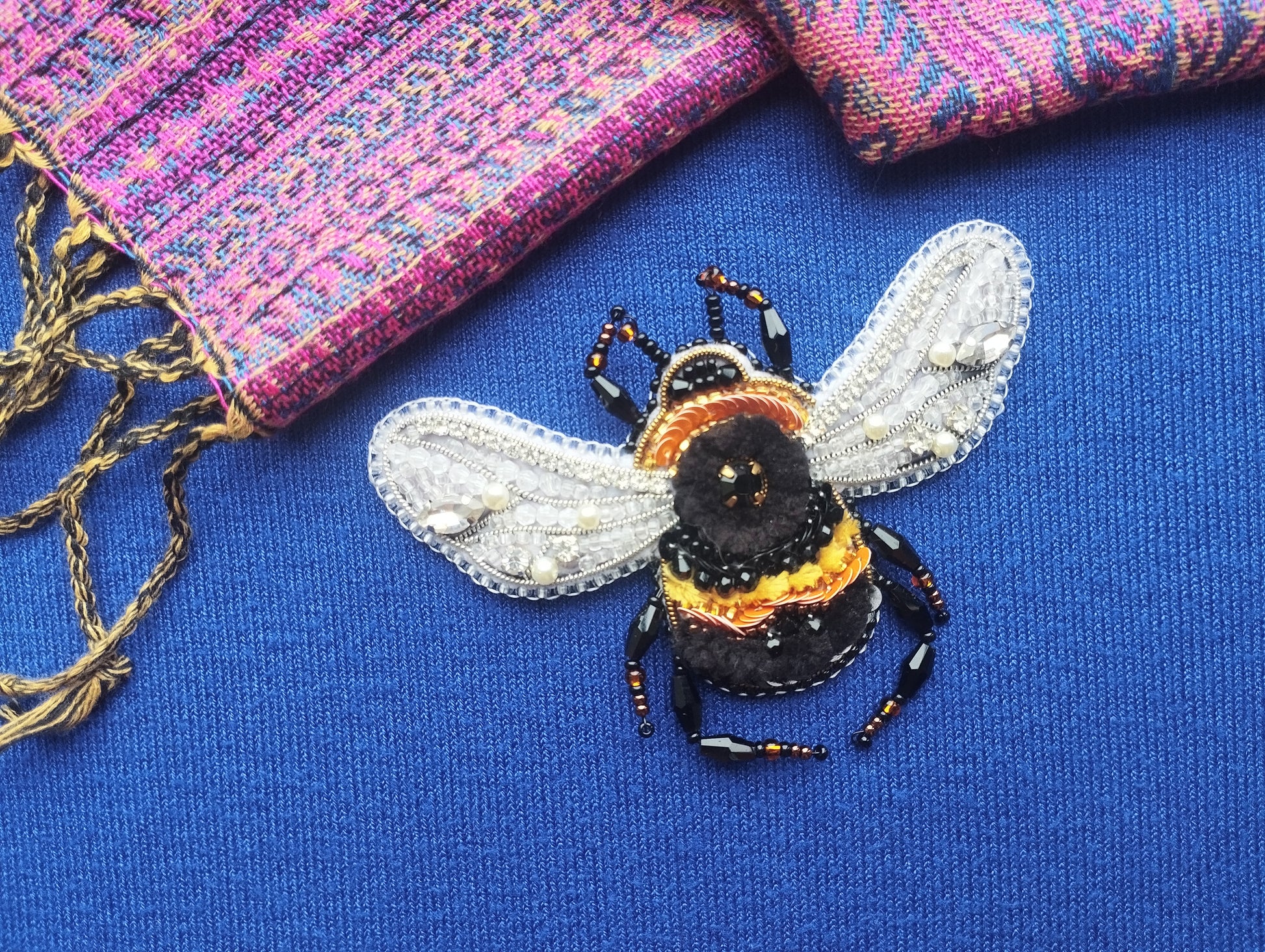 a detailed embroidered bee on a blue fabric surface, with a pink and purple patterned fabric visible in the background.