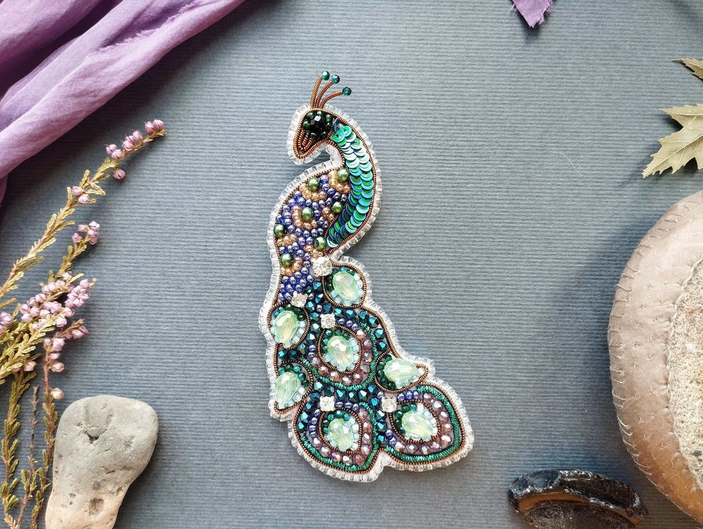 a decorative peacock brooch with a vibrant and intricate design, surrounded by various natural elements such as flowers, leaves, and rocks.