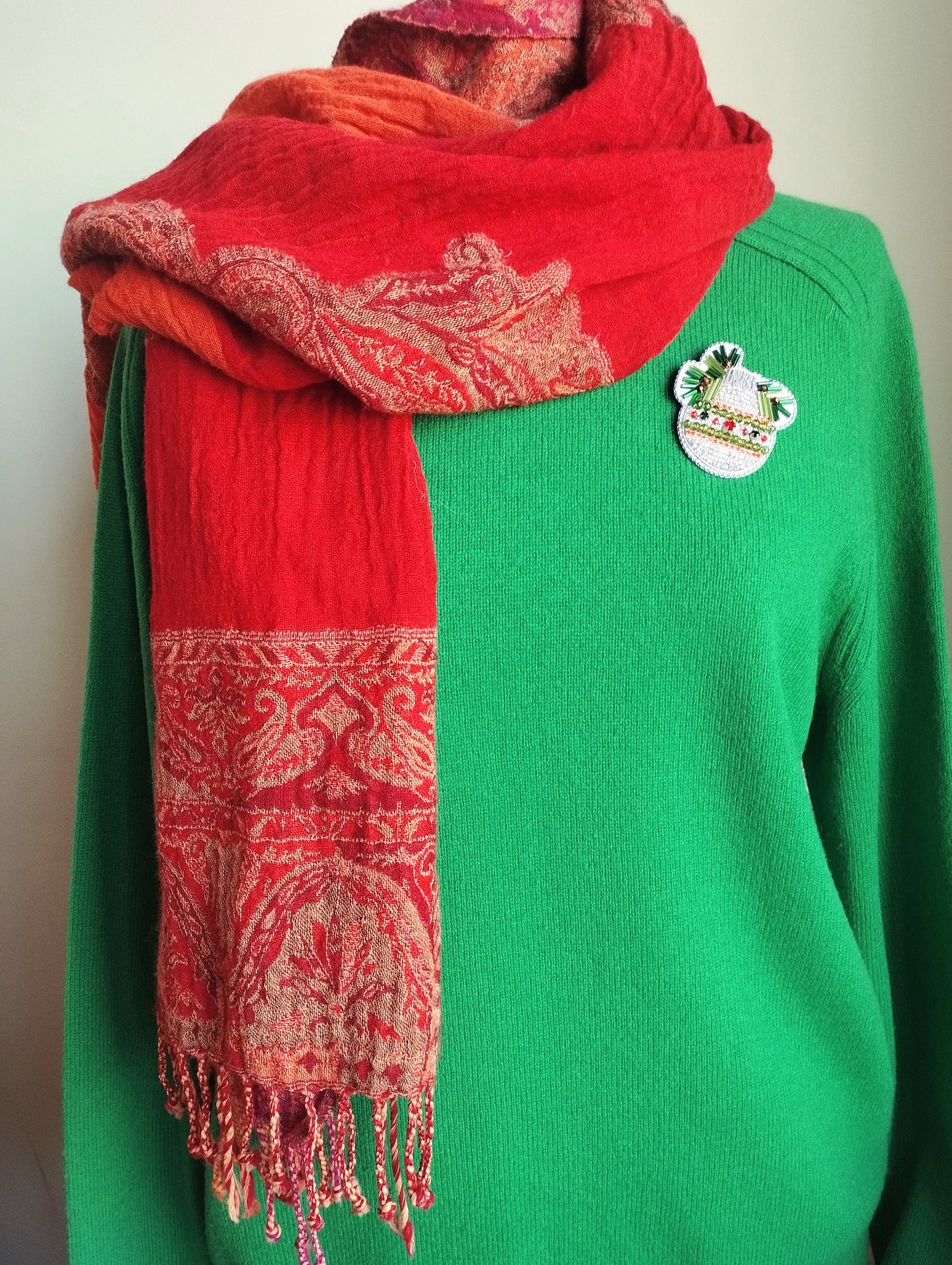 A mannequin is wearing a green sweater with a red and orange patterned scarf draped over it. The scarf has fringe details and a small pin on the sweater.