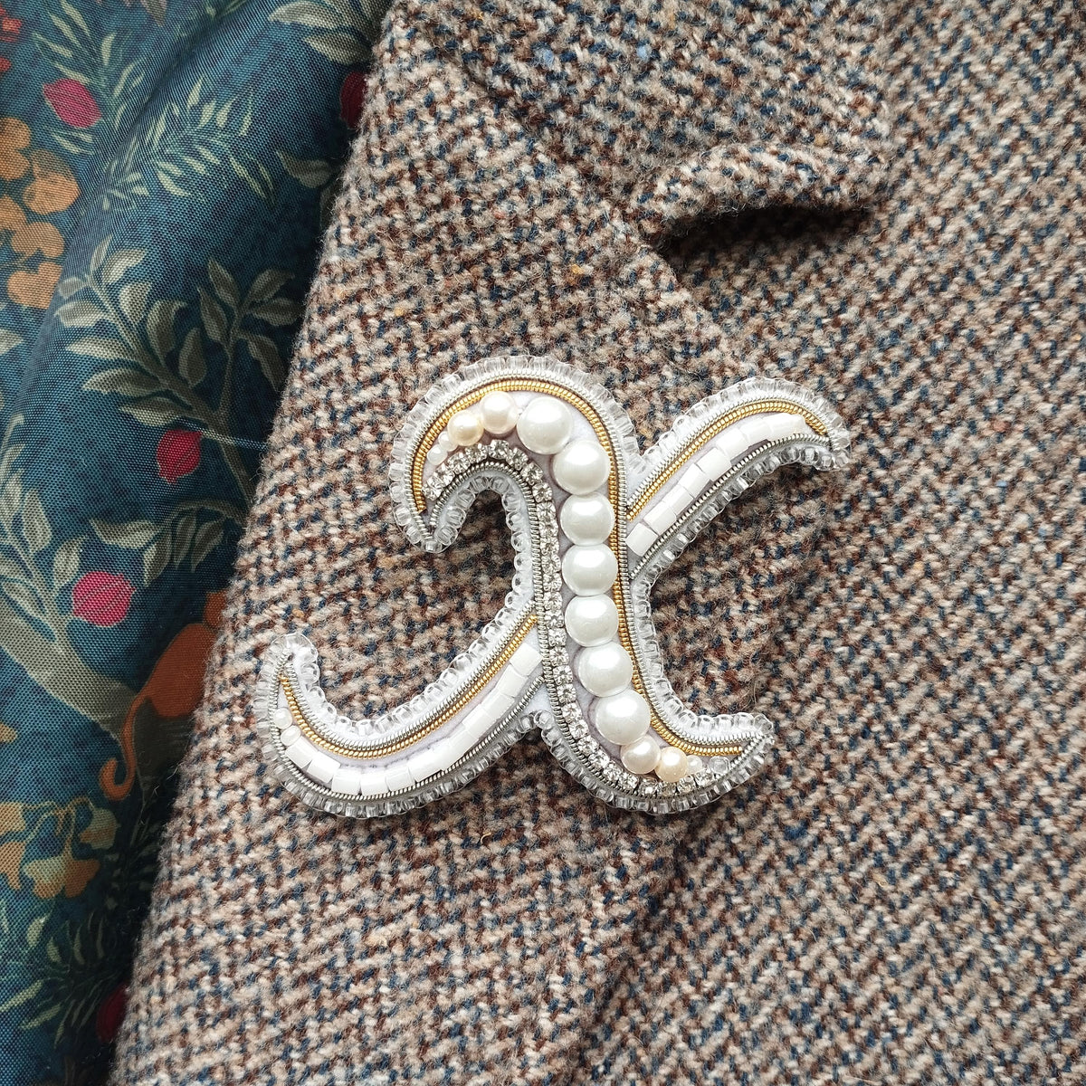 A close-up image of a decorative monogram brooch with pearls and a metallic sheen, placed on a tweed jacket.