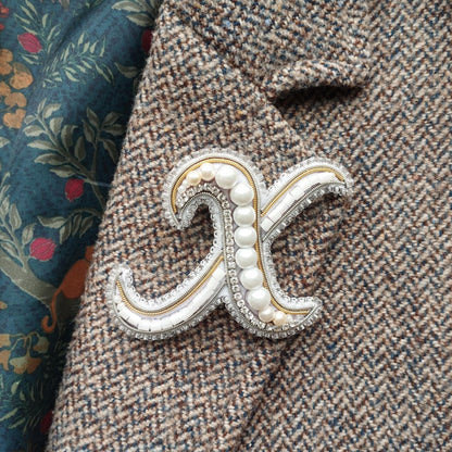 A close-up image of a decorative monogram brooch with pearls and a metallic sheen, placed on a tweed jacket.
