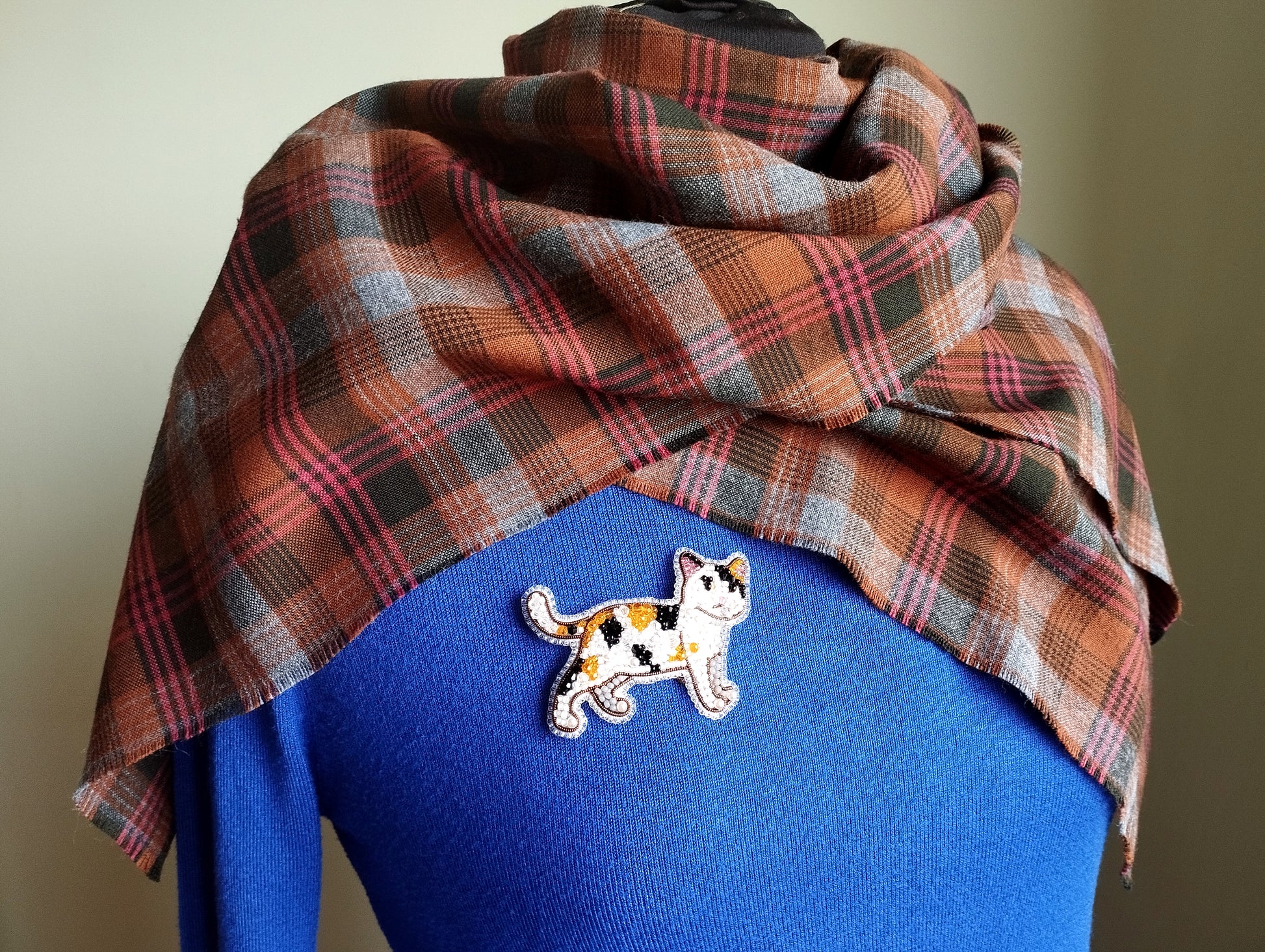 A plaid scarf is draped over a blue sweater, with a small embroidered cat pin or badge pinned to the sweater.