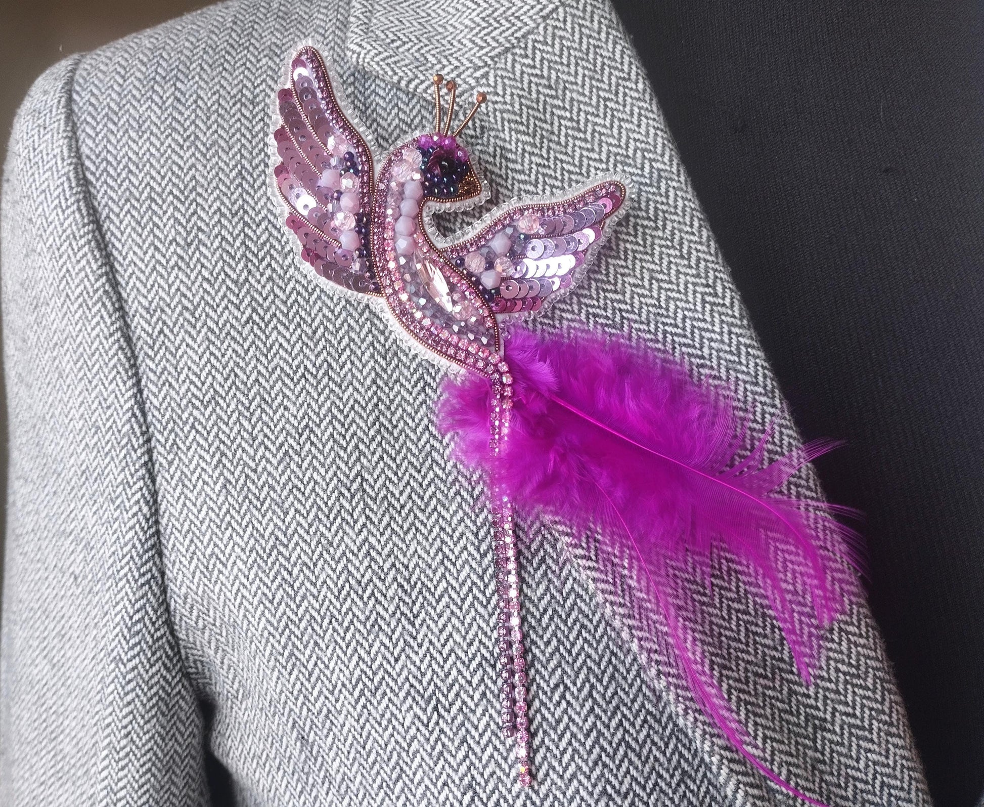 A close-up of a person wearing a gray tweed jacket with a decorative pin featuring a pink bird and a feather attached to the lapel.