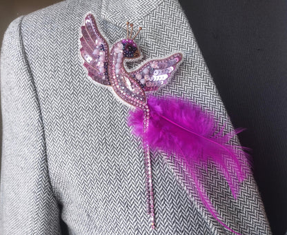 A close-up of a person wearing a gray tweed jacket with a decorative pin featuring a pink bird and a feather attached to the lapel.