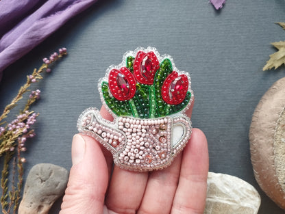 A hand holding a decorative beaded object resembling a teapot with red flowers, against a gray background with various natural elements such as leaves and rocks.